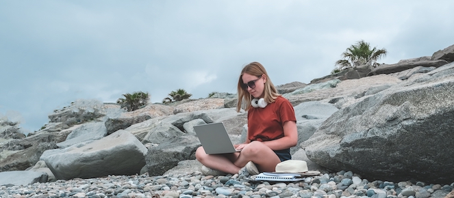 Woman wearing headphones and sun glasses outside on the rocks working on her laptop in a work life balance