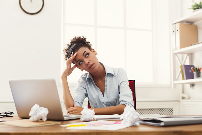 Deadline stress concept - sad african-american business woman sitting at desktop in office and holding hand on head. Hard working day.