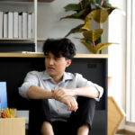 Asian man with his office packed in a box, sitting on the floor in front of his desk after being laid off