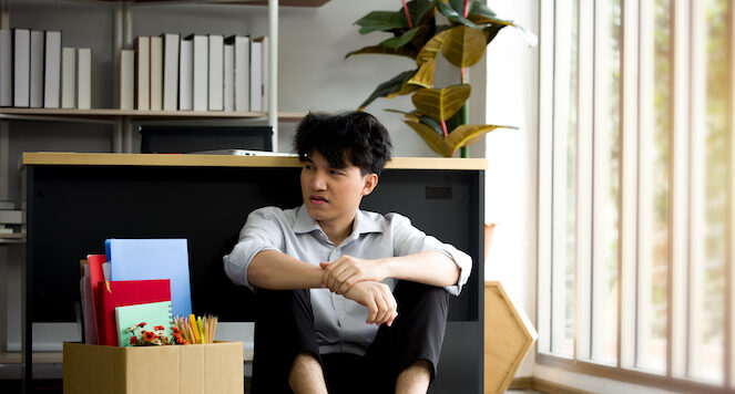 Asian man with his office packed in a box, sitting on the floor in front of his desk after being laid off