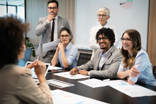 A group of diverse employees engaged with their leader in conversation at a board table.