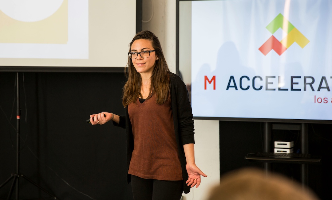 Women speaking on a stage with her pitch deck behind her, pitching her business. 
