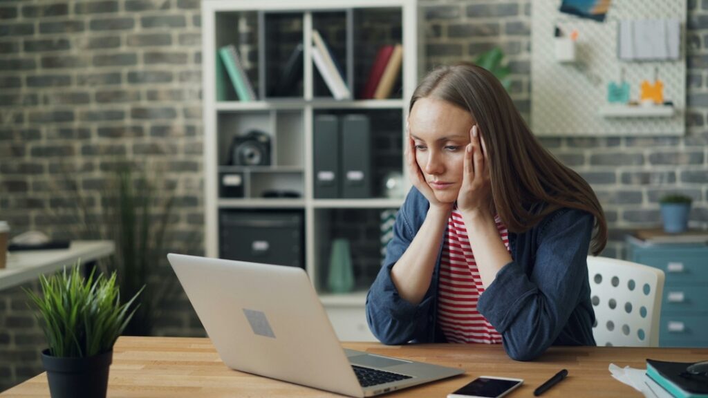 Overwhelmed, stressed woman sitting at her desk with her hands on the side of her face staring at a laptop.