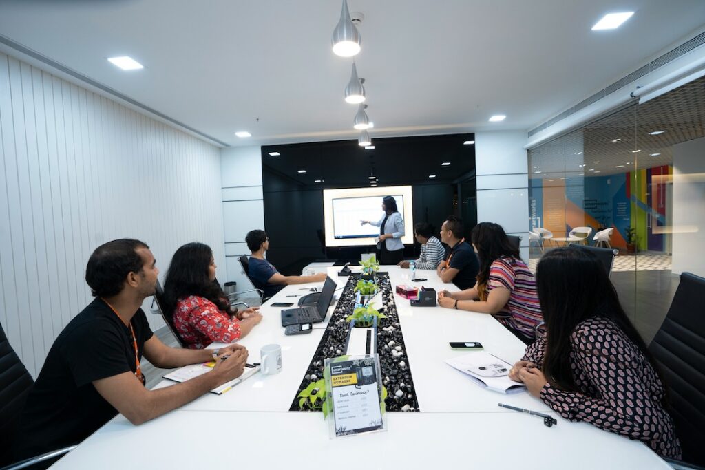 A team comfortably sitting at a table with a leader up front talking and using a whiteboard to demonstrate a point. 