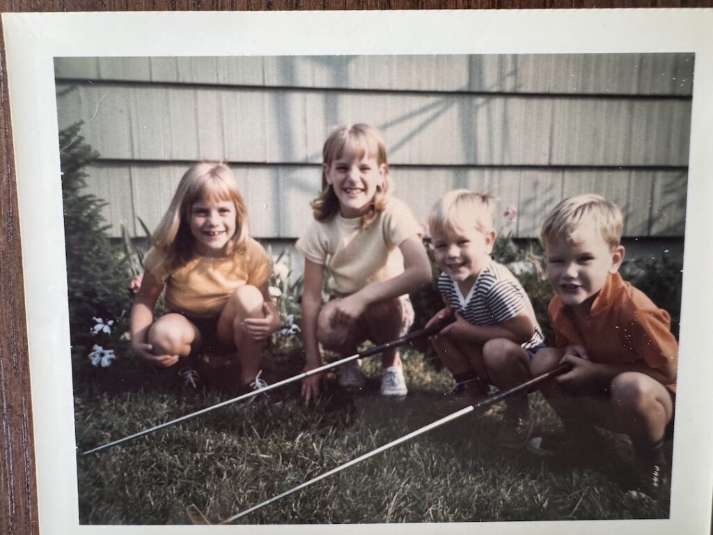 Four siblings squatting in the grass smiling for the camera shot and two are holding golf clubs.