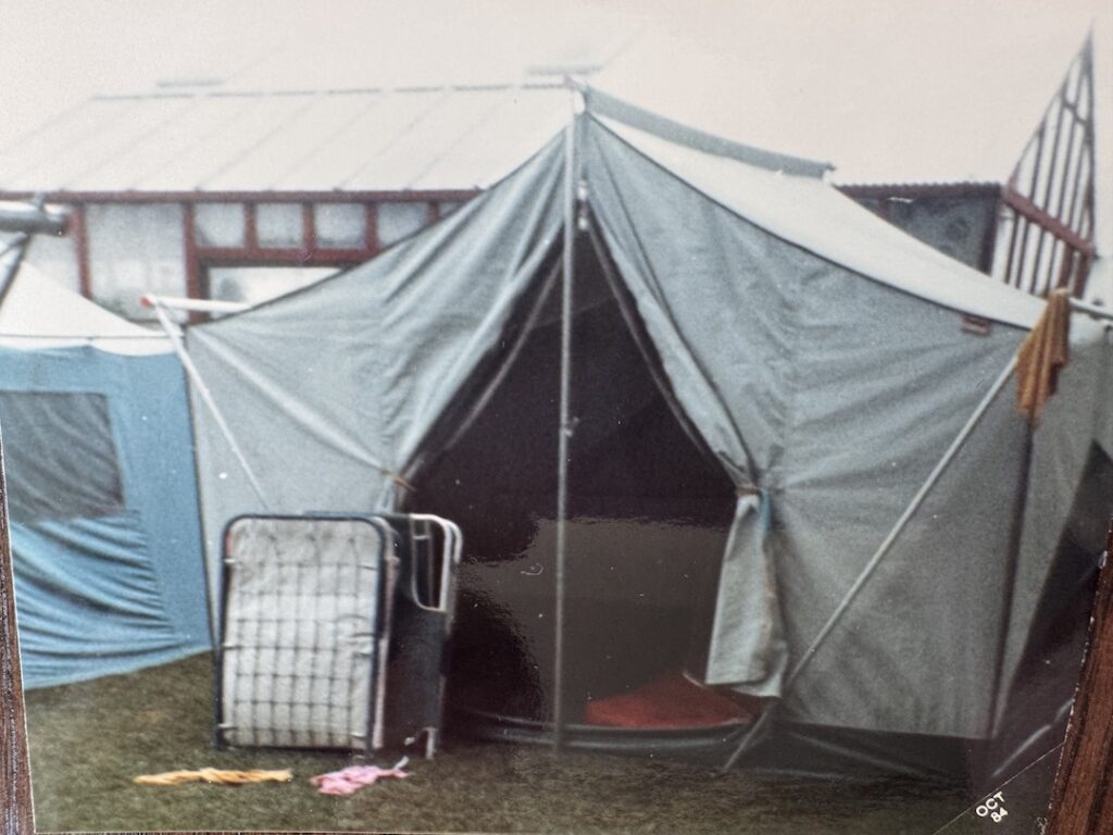 A blue canvas tent with the doors flapped open and two cots leaning against it.