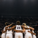 A diverse team huddle of a women's basketball team in the arena with a large crowd in the background.
