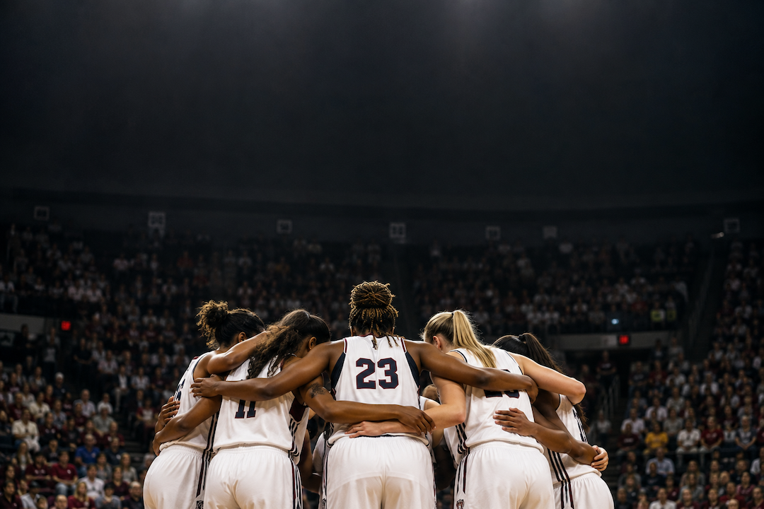 A diverse team huddle of a women's basketball team in the arena with a large crowd in the background.