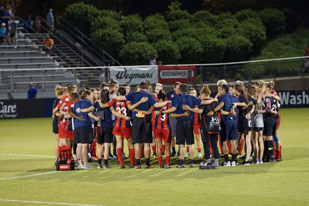 Entire soccer team and coaches in a huddle in the middle of the field with arms around each other. 